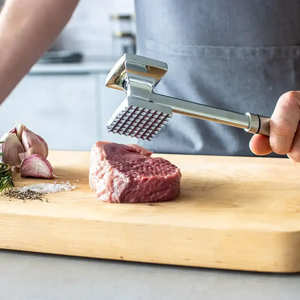 Person using a meat mallet on a piece of meat on a wooden cutting board with garlic and herbs.
