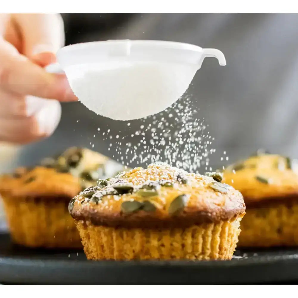 Person dusting powdered sugar over a muffin with pumpkin seeds on a dark surface.