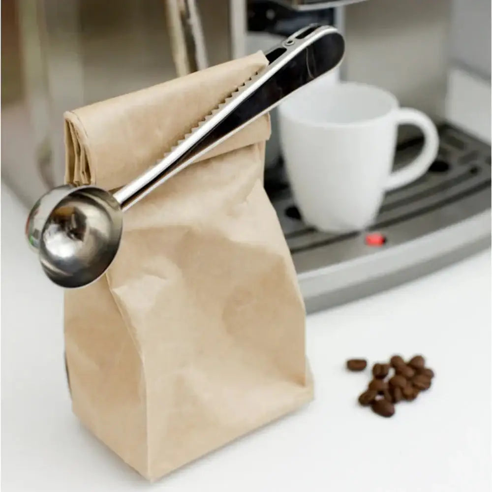 Coffee scoop lifting a brown paper bag of coffee beans with a white cup and coffee beans on a counter.