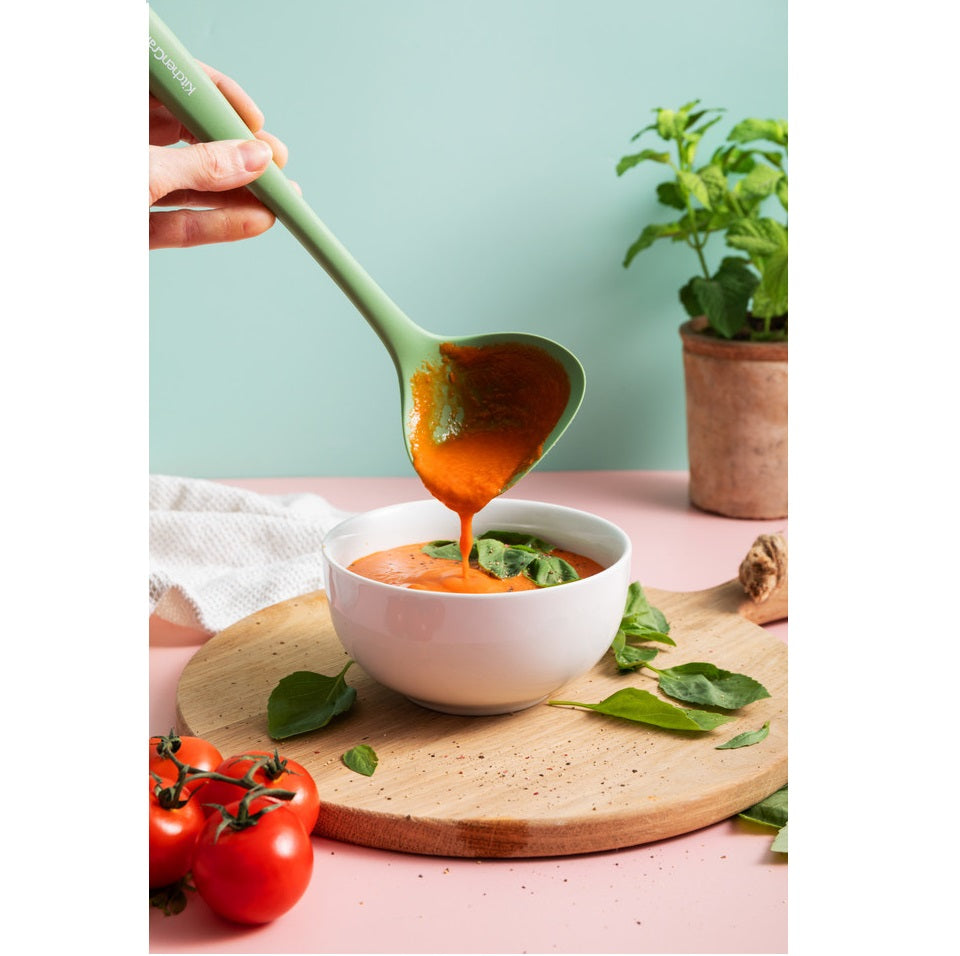 Serving tomato-based soup from a ladle into a white bowl on a pink surface with a plant in the background.