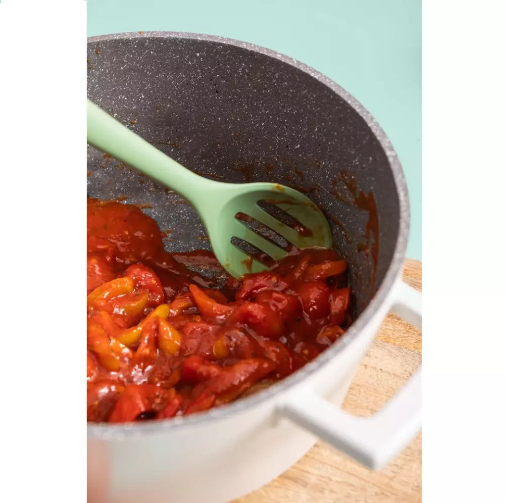 Potted dish with sautéed vegetables and a green slotted spoon on a light blue background