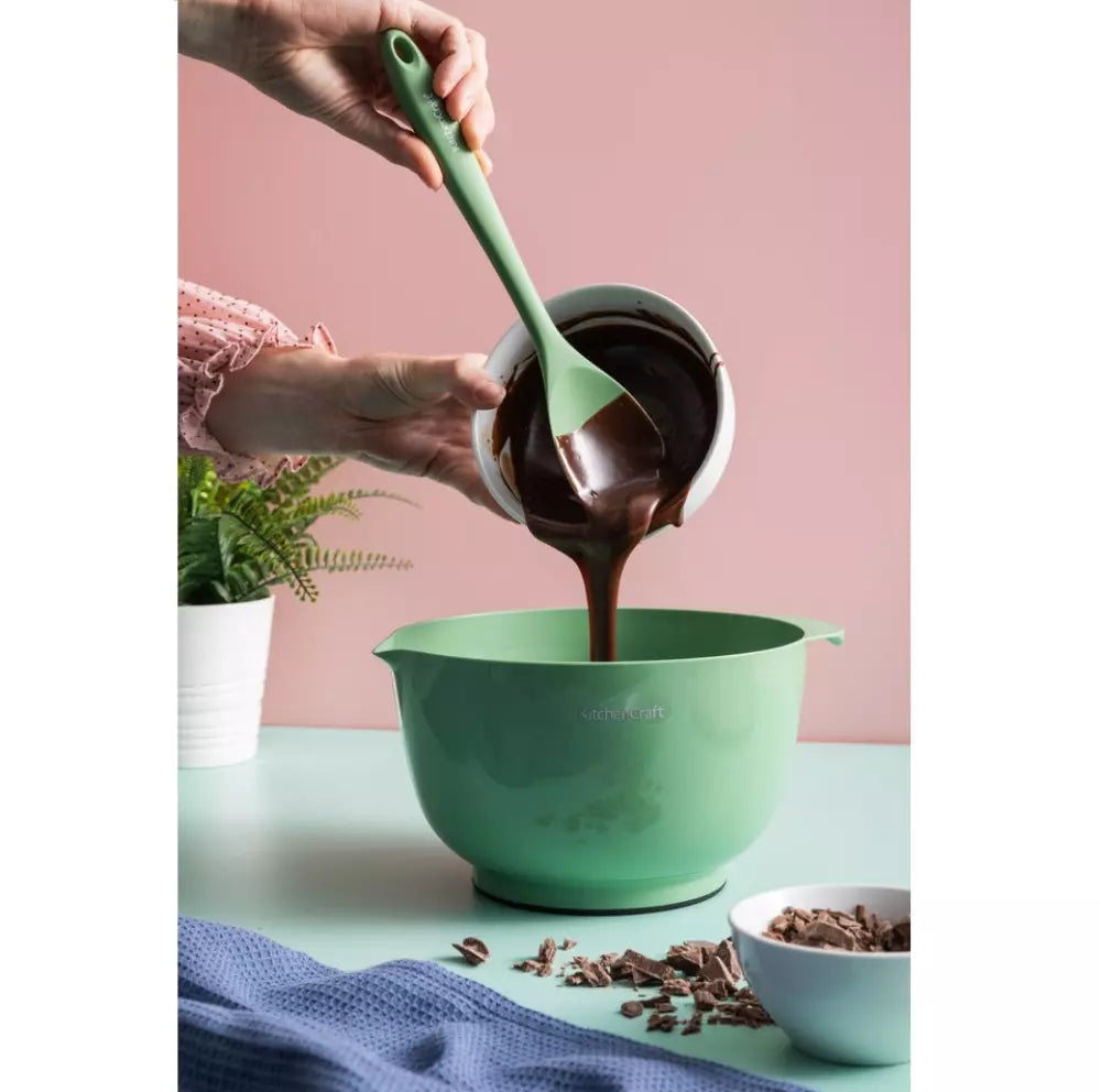 Person using a green spatula to transfer chocolate from a small bowl into a larger green mixing bowl on a pink background.