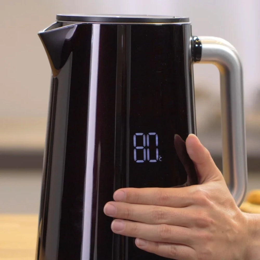 Black electric kettle with digital temperature display held by a hand on a blurred kitchen background