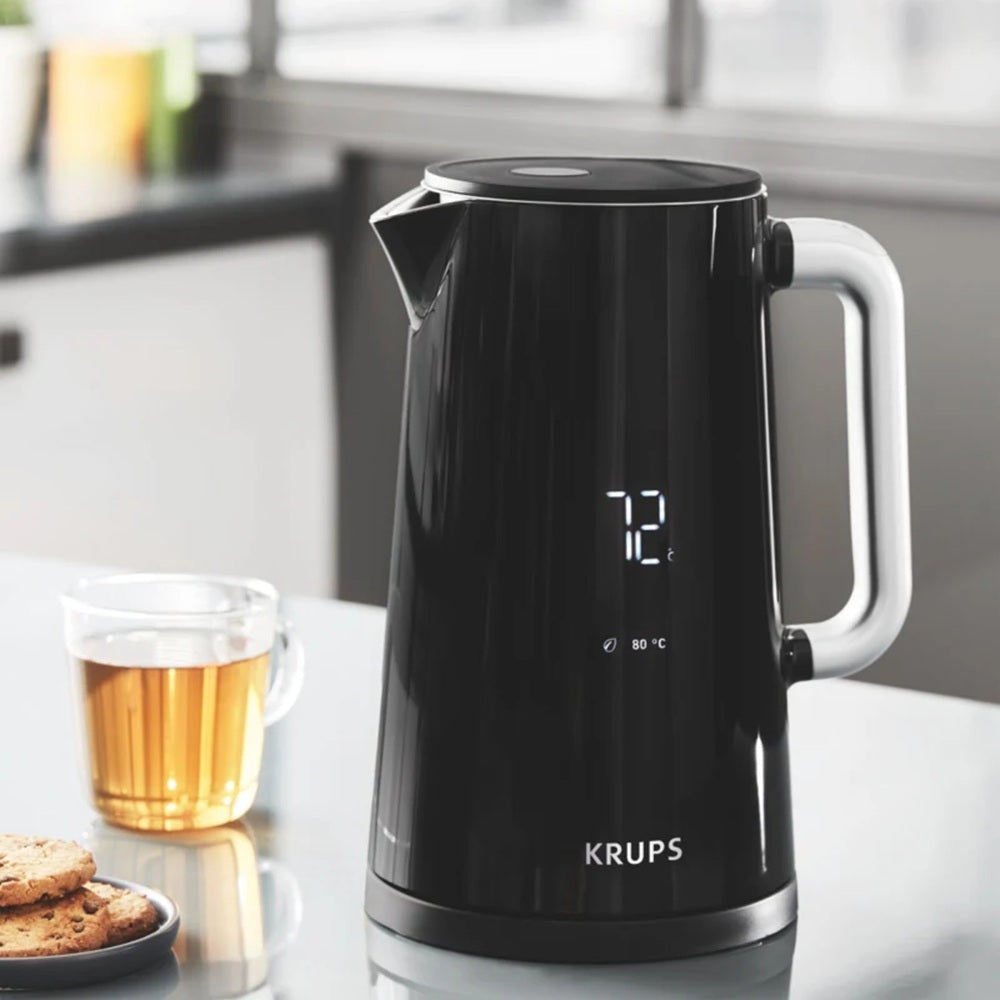 Black Krups electric kettle on a kitchen counter with a glass of tea and cookies in the background.