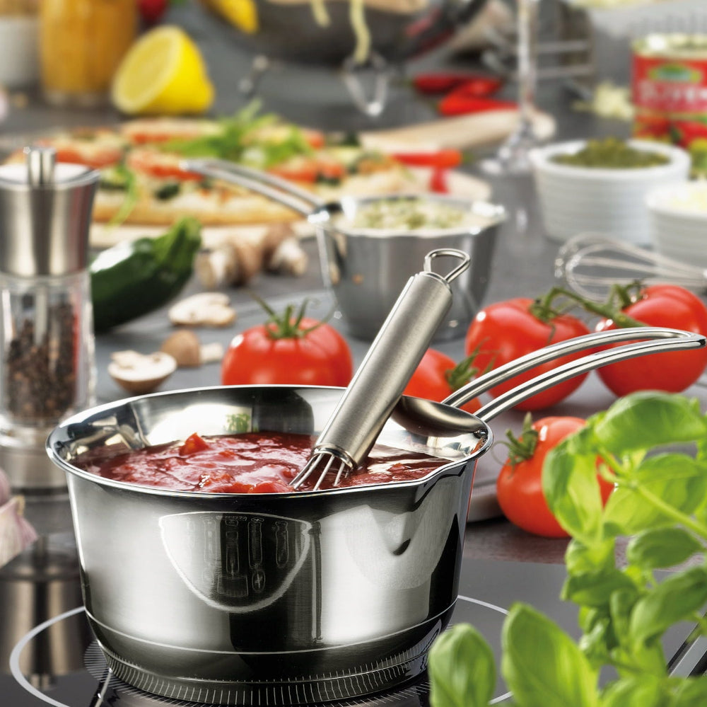 Stainless steel pot with a ladle on a kitchen counter with vegetables and cookware in the background