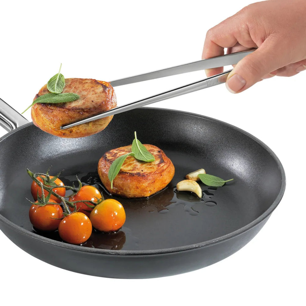 A black non-stick frying pan on a white background, being held by a hand with metal tongs. Inside the pan are two golden-brown cakes topped with green herbs, a few cherry tomatoes on the vine, and garlic cloves.
