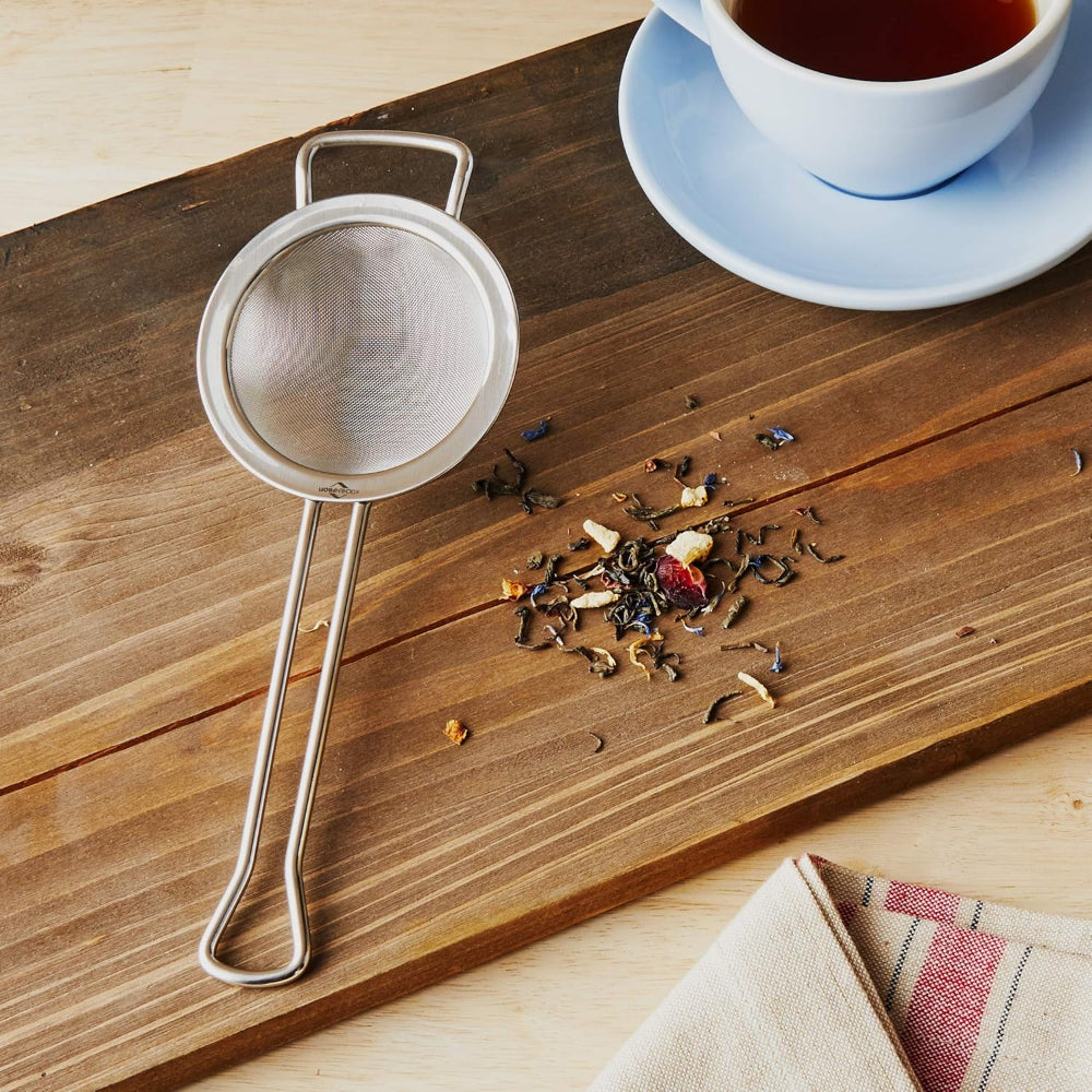 Tea strainer on a wooden surface with tea leaves and a cup of tea in the background.
