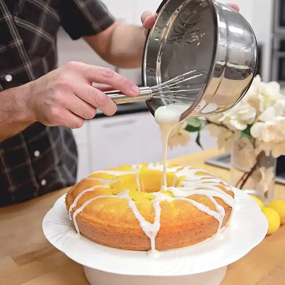 Person pouring a white glaze over a cake with a whisk on a kitchen counter.