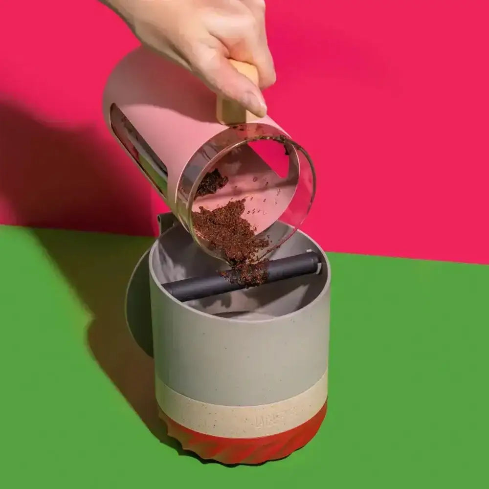 Hand using a pink and clear coffee grinder to pour coffee grounds into a container on a green and pink background.