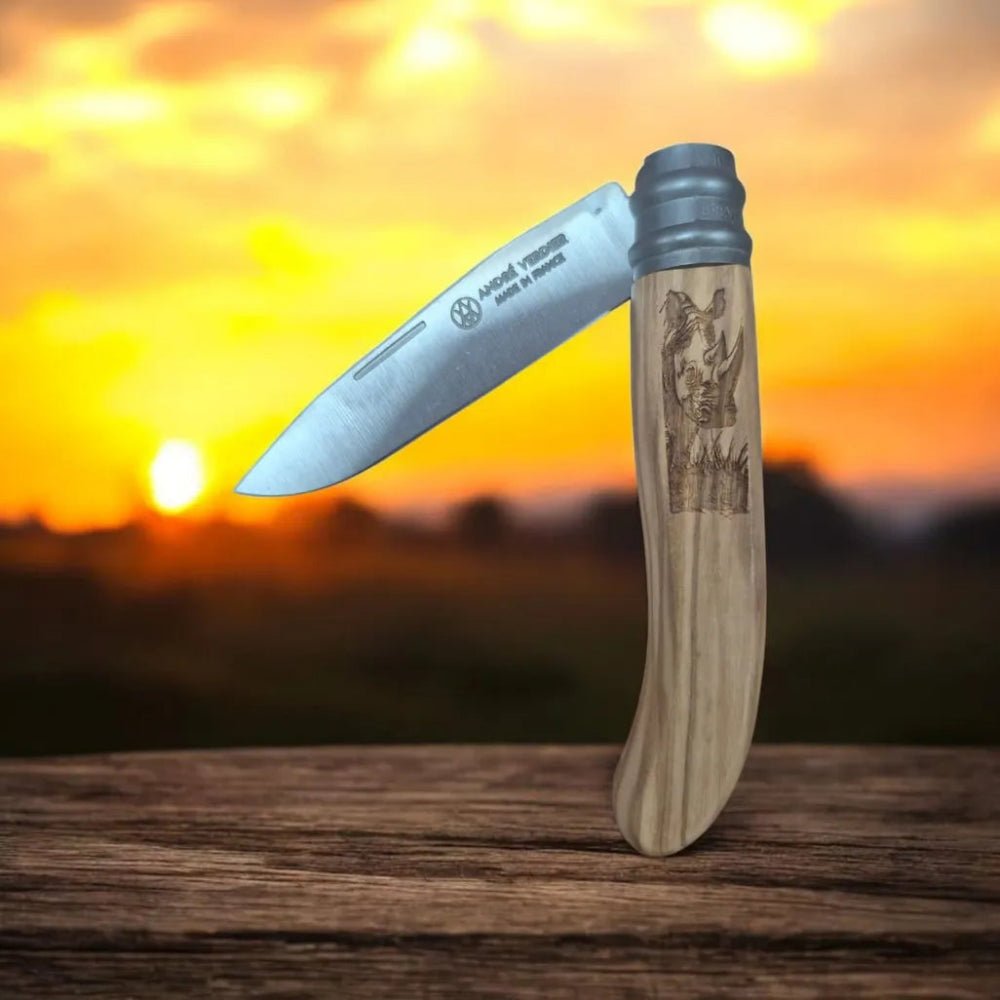A folding pocket knife with a silver steel blade and a light wood handle featuring an engraved rhinoceros design, standing upright on a wooden surface against a sunset background.