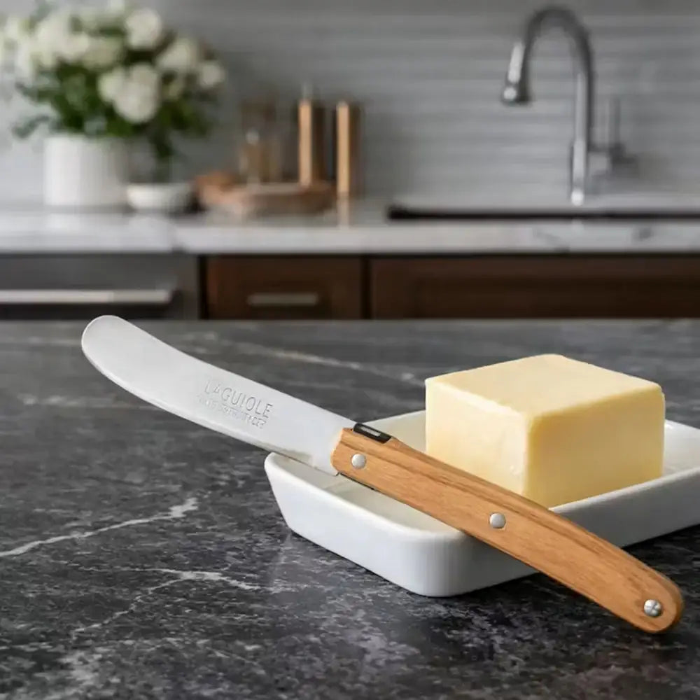 Butter knife with wooden handle and white blade on a butter dish with a stick of butter, set against a kitchen counter.