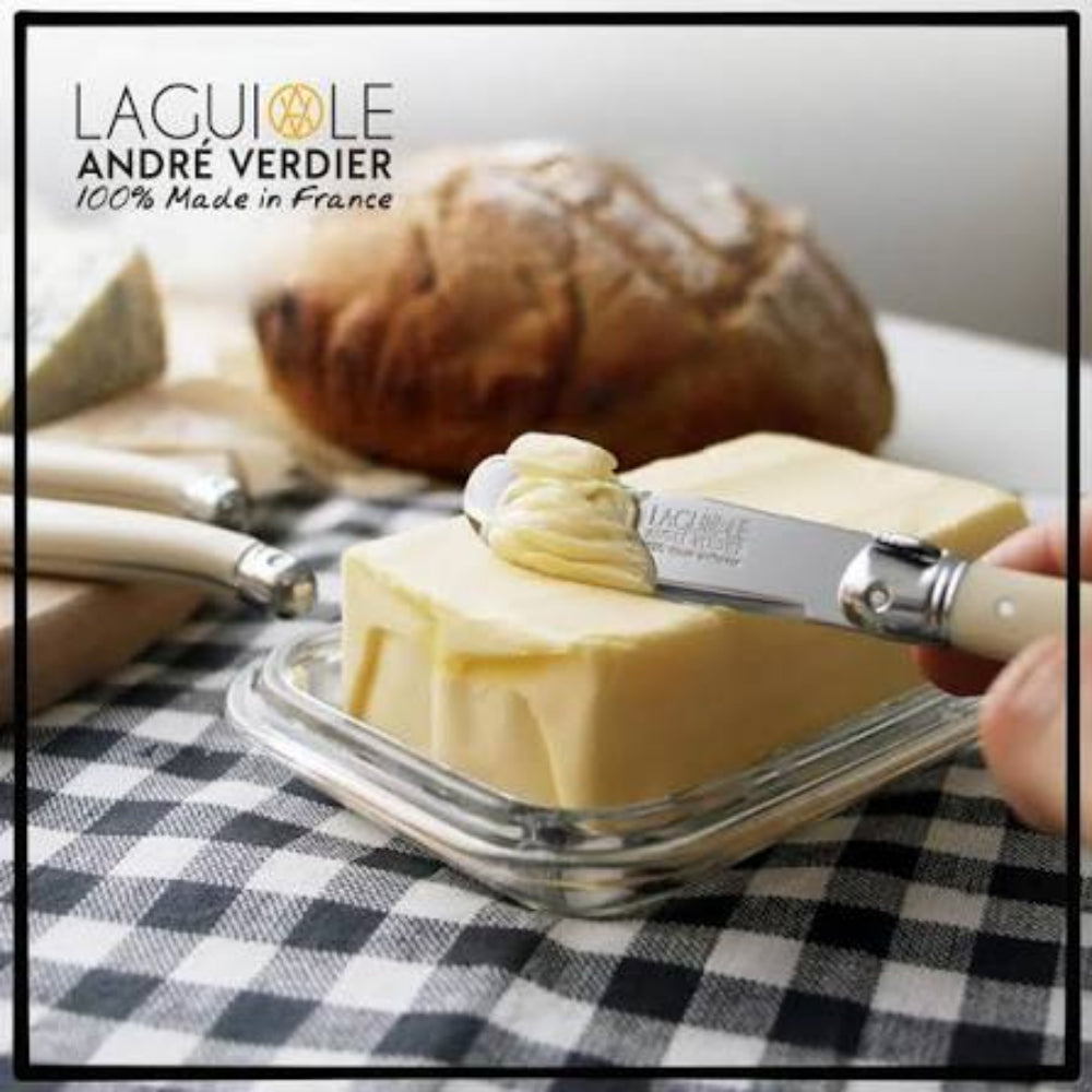 A close-up of a hand using a Laguiole André Verdier butter knife to spread butter on a stick in a glass dish, with bread and additional knives in the background on a black-and-white checkered tablecloth. The Laguiole André Verdier logo appears in the top left.