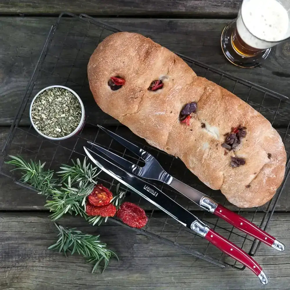 Loaf of bread with dried fruits, a small bowl of herbs, and two knives on a wooden surface.