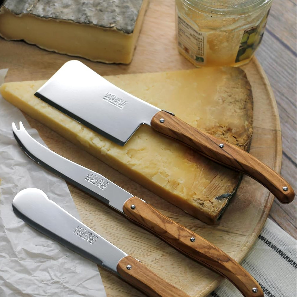 Three cheese knives with light brown wooden handles and silver stainless steel blades are arranged on a round wooden board with various cheeses, including blue cheese and a wedge of yellow cheese, alongside a slice of bread and a jar of spread.
