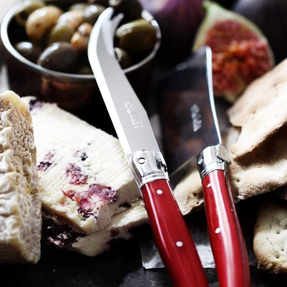 Two red-handled knives on a plate with bread and olives in the background