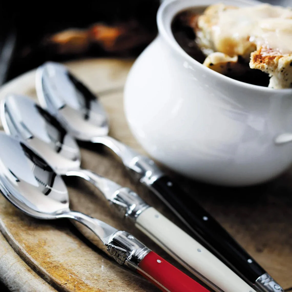 Set of silver spoons with red and black handles on a wooden surface next to a bowl of soup.