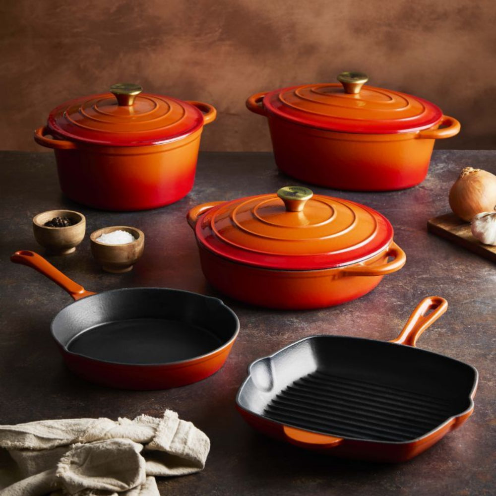 A set of cast iron cookware in a vibrant orange-red gradient finish, including three lidded pots, a skillet, and a grill pan, arranged on a dark rustic tabletop with small food props.
