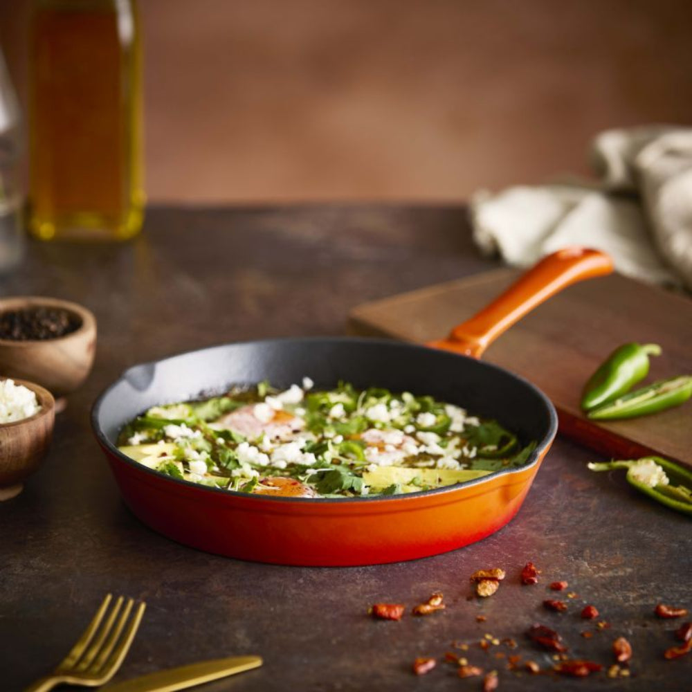 A red and black cast iron skillet filled with a green and white breakfast dish, set on a rustic dark wooden table with a wooden cutting board, green peppers, and spices.