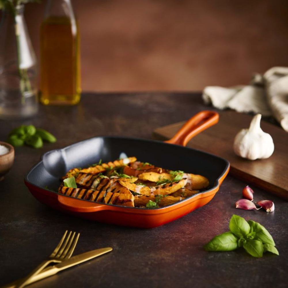 An orange and black cast iron skillet filled with grilled potatoes and greens, styled on a dark rustic table with garlic, basil, a bottle of oil, and gold cutlery.
