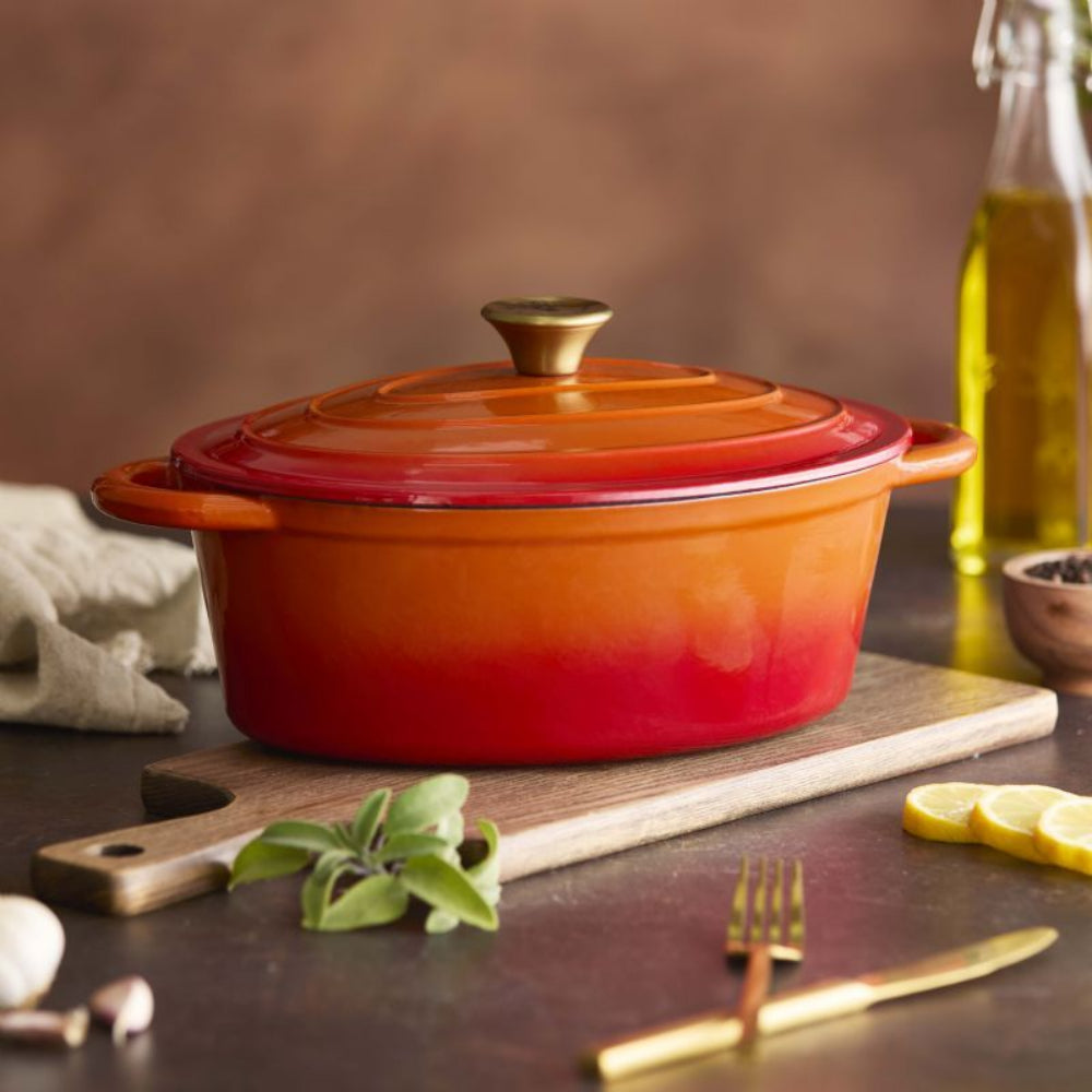An orange-to-red gradient enameled cast iron Dutch oven with a matching lid and gold-toned knob, styled on a wooden cutting board with garlic, sage, lemon slices, a fork, and a knife on a dark table.
