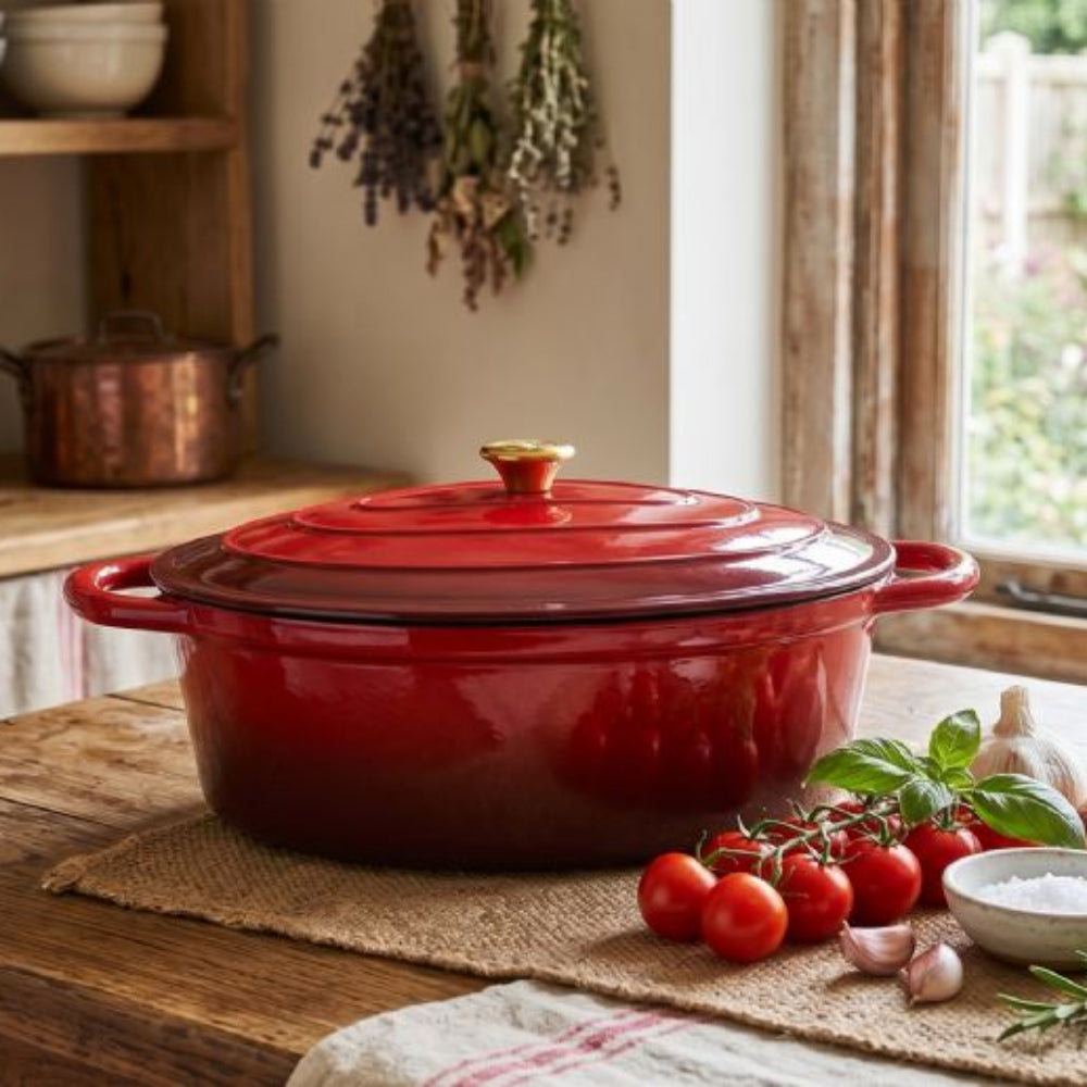 Red cast iron casserole dish on a wooden table with tomatoes and herbs in a kitchen setting.