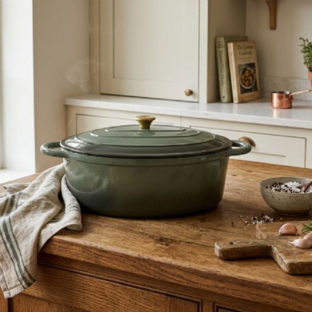 Green cast iron cookware on a wooden kitchen counter with ingredients and books in the background.