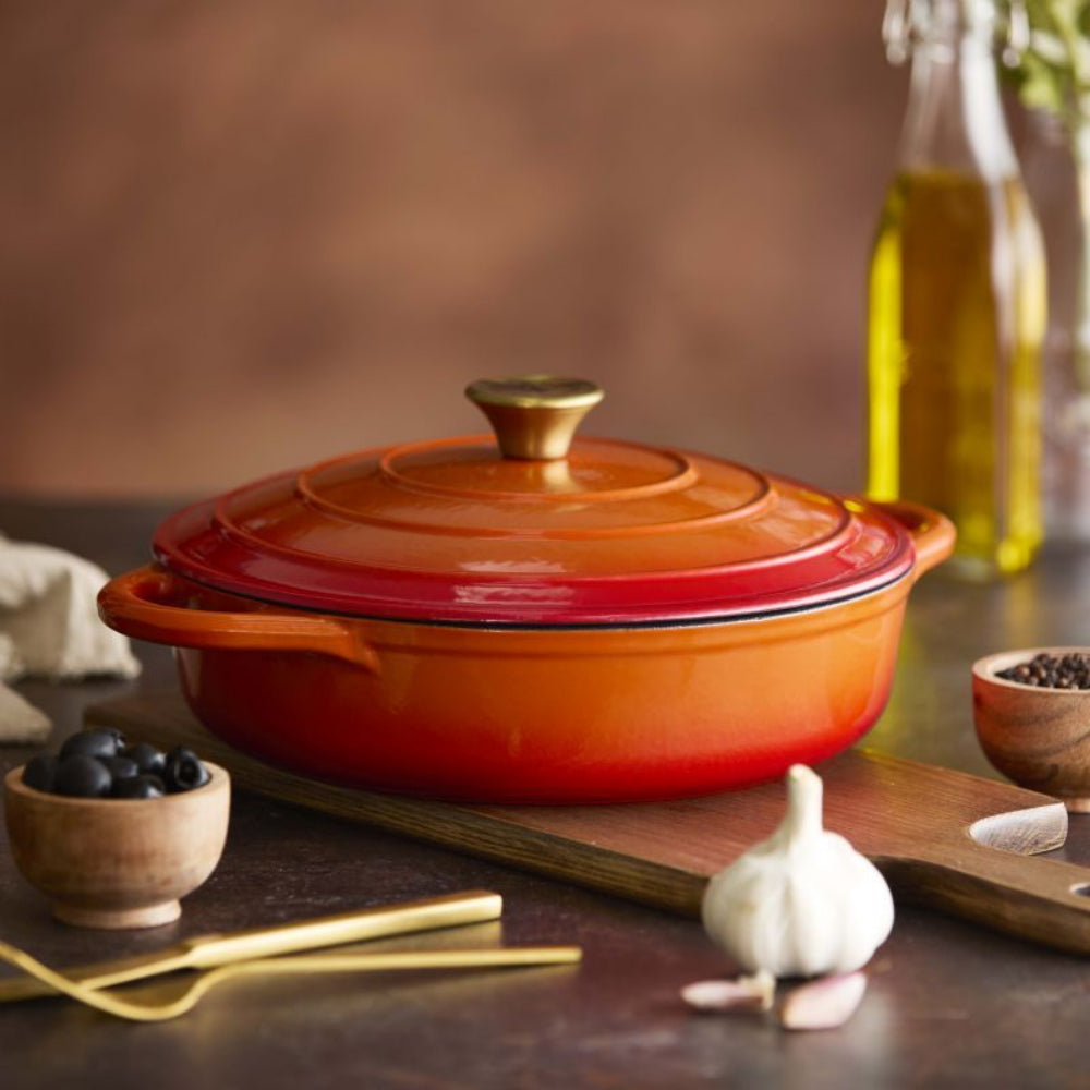 An orange and red gradient enameled cast iron Dutch oven with a matching lid, styled on a wooden cutting board with garlic, olives, and a bottle of olive oil in the background.