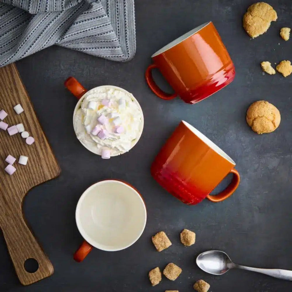 Two orange mugs with a white mug on a dark surface with cookies and marshmallows.