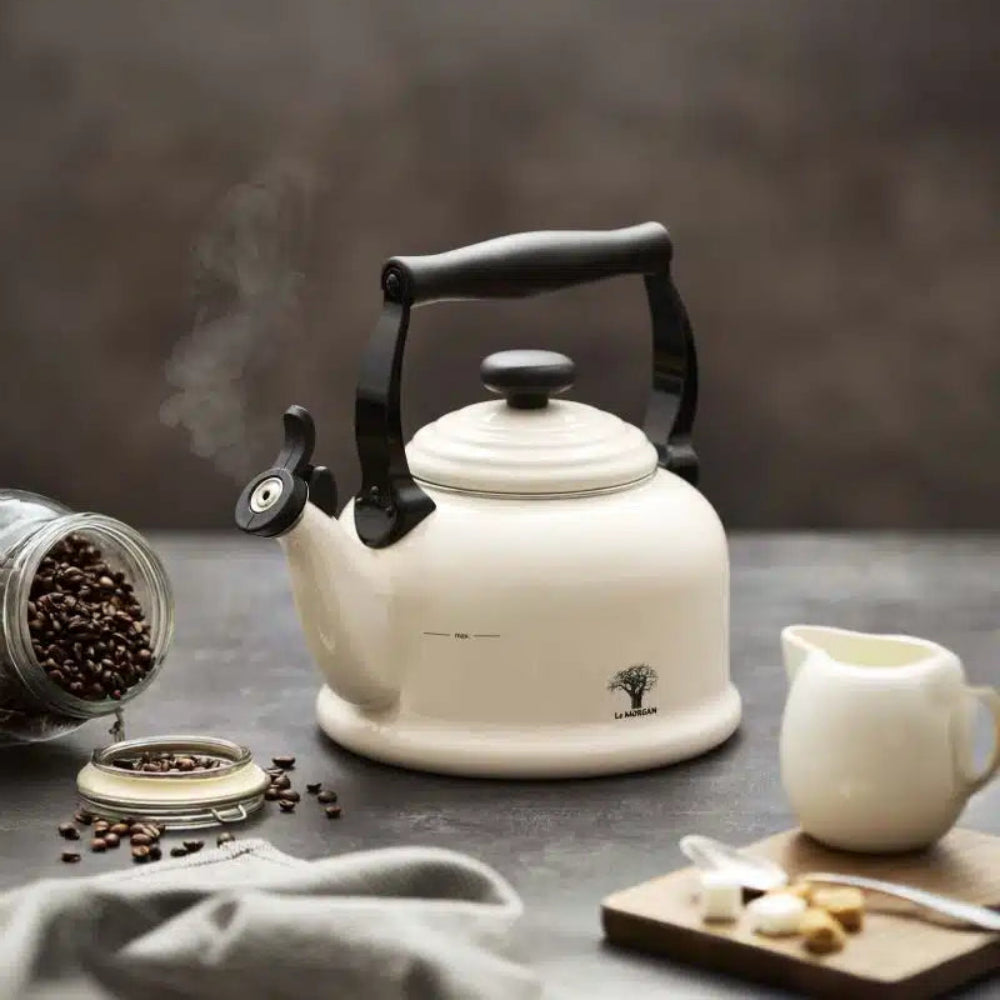 A cream-colored enamel kettle with a black handle and spout, shown on a dark tabletop with coffee beans, a small cream pitcher, and steam rising. The kettle has a small black logo near the base.