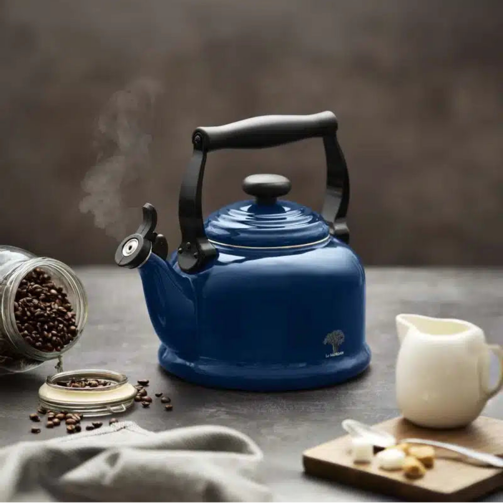 A blue enamel tea kettle with a black handle sits on a dark tabletop with steam rising. Coffee beans spill from a jar, with a white creamer pitcher and a small cutting board nearby against a dark gray background.