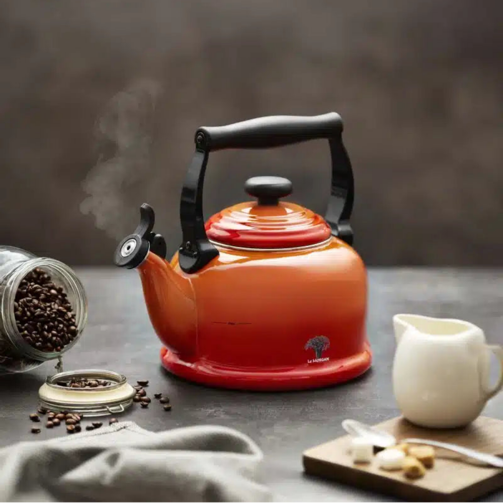 An orange enamel tea kettle with a black handle and lid knob, shown on a dark tabletop with coffee beans, a white creamer, and steam rising. The kettle has a small tree logo near the base.