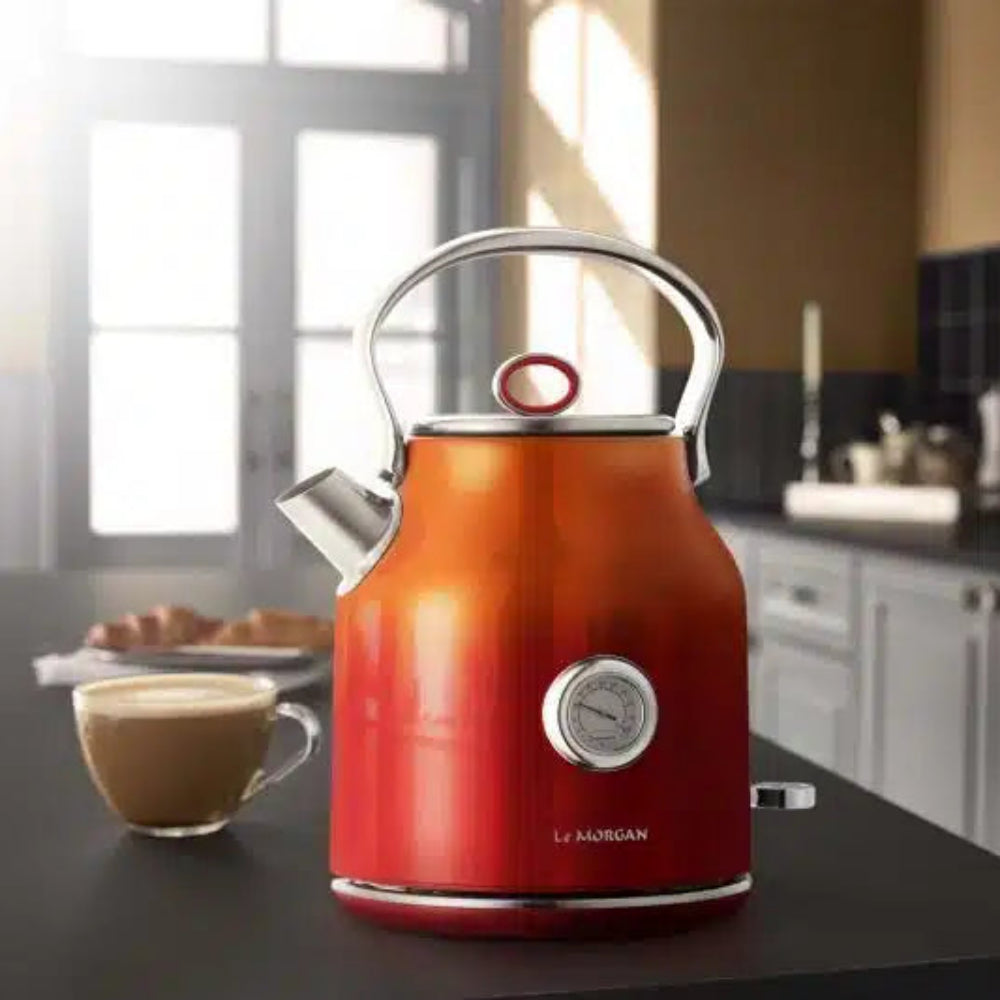 A Le Morgan electric kettle with a red-to-orange gradient finish, set on a dark kitchen counter next to a cup of coffee.