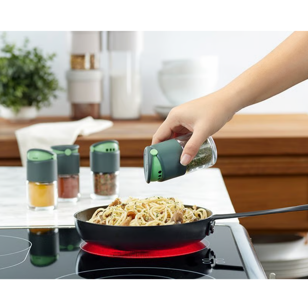 A hand sprinkles seasoning from a green and clear spice shaker onto pasta in a black frying pan on a stovetop. Three matching spice shakers sit on the counter in a bright kitchen setting.