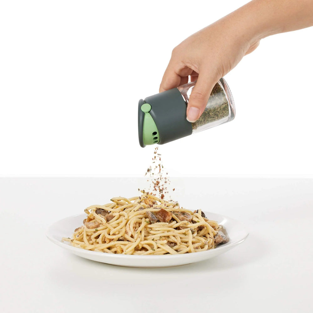 Hand sprinkling herbs from a small container onto a plate of pasta.