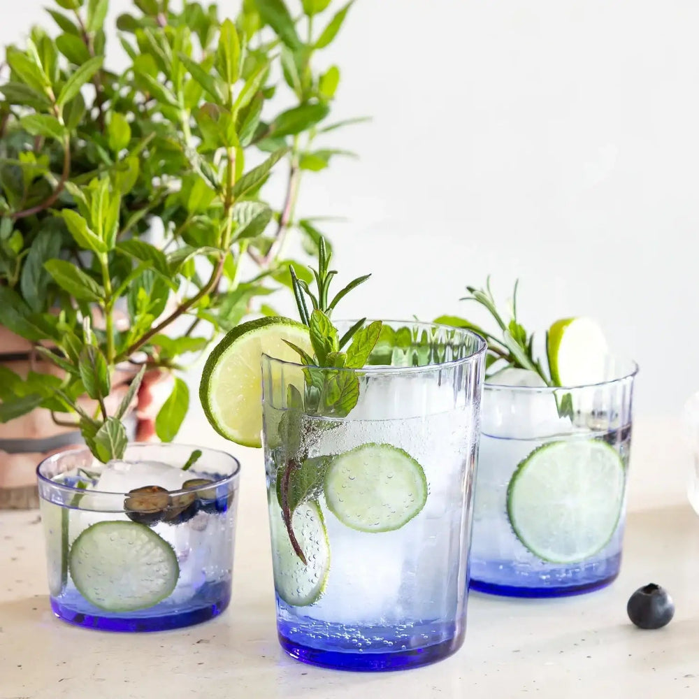 Three glasses of blue liquid with lime and mint on a white surface with plants in the background