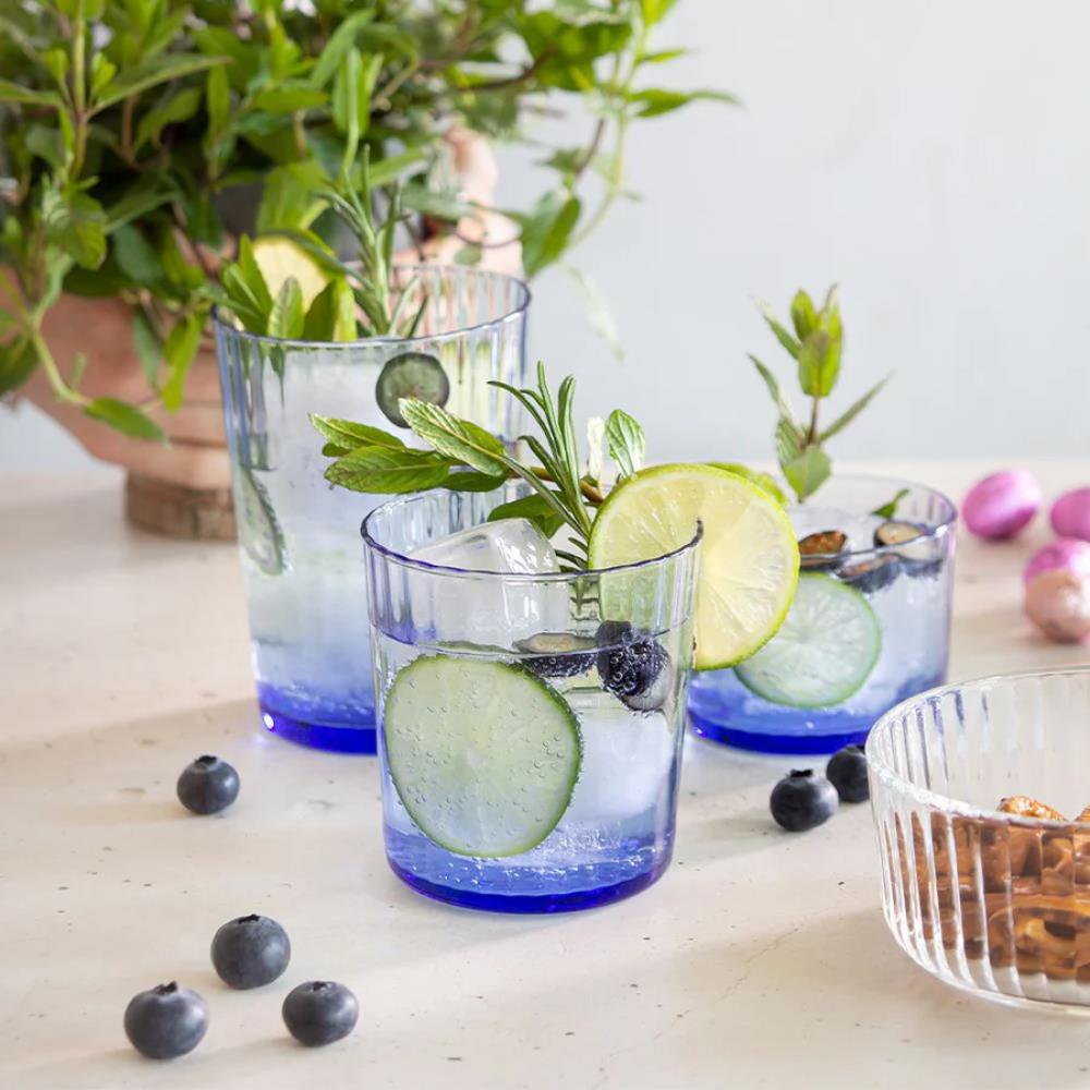 Three glasses of water with cucumber slices, lime, and blueberries on a wooden surface with plants in the background.