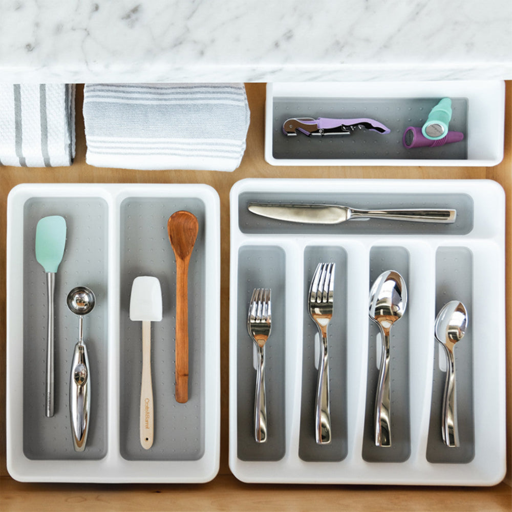 White and grey cutlery trays with silverware and utensils placed inside a wooden drawer.