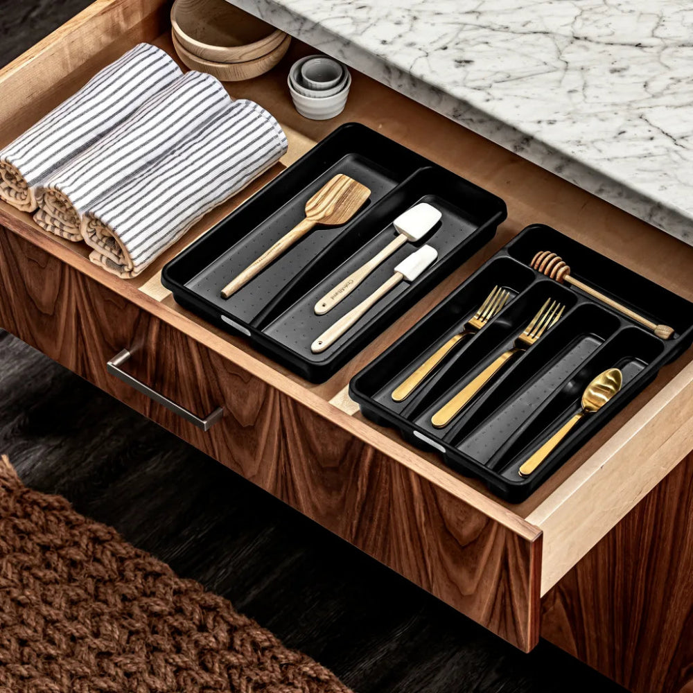 Wooden drawer with black trays containing kitchen utensils on a marble countertop.