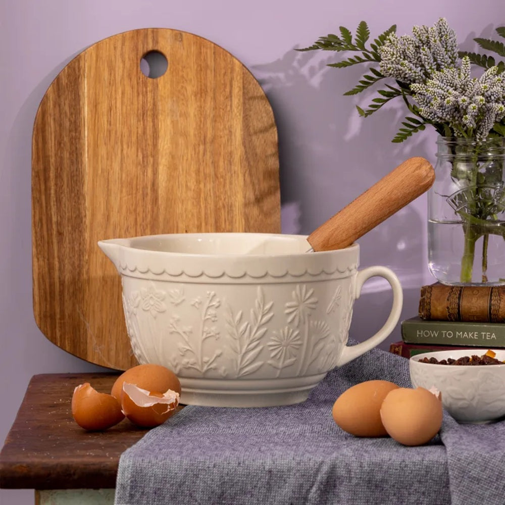 White ceramic mixing bowl with wooden handle on a kitchen counter with eggs and a cutting board.