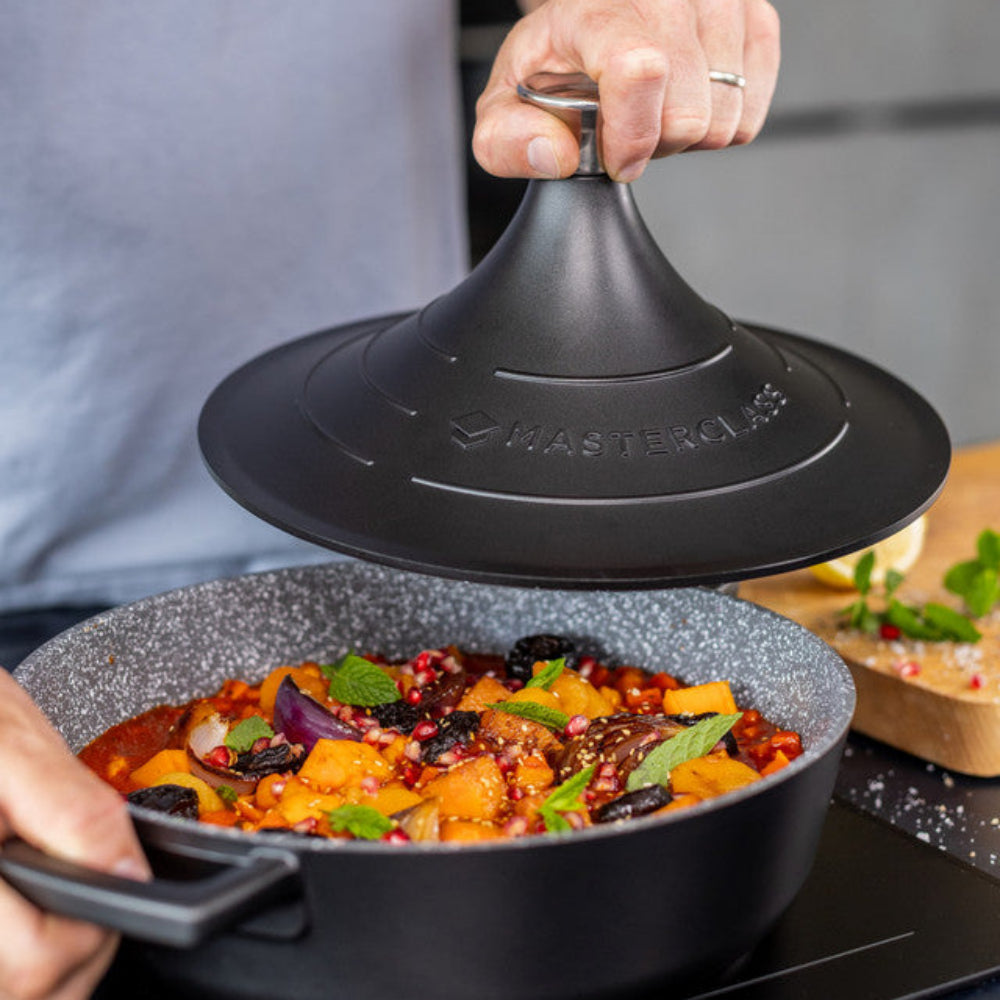 Person lifting a black tagine over a pan of food on a kitchen counter.