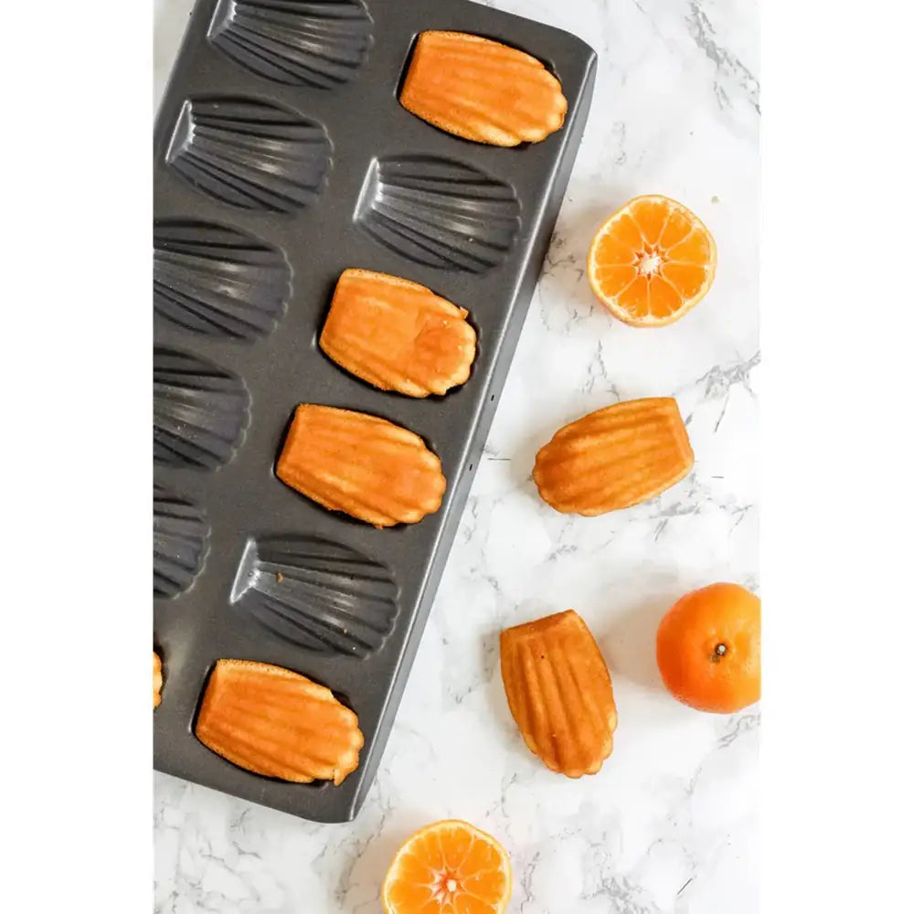 Madeleine cookie mold with baked cookies and sliced oranges on a marble surface