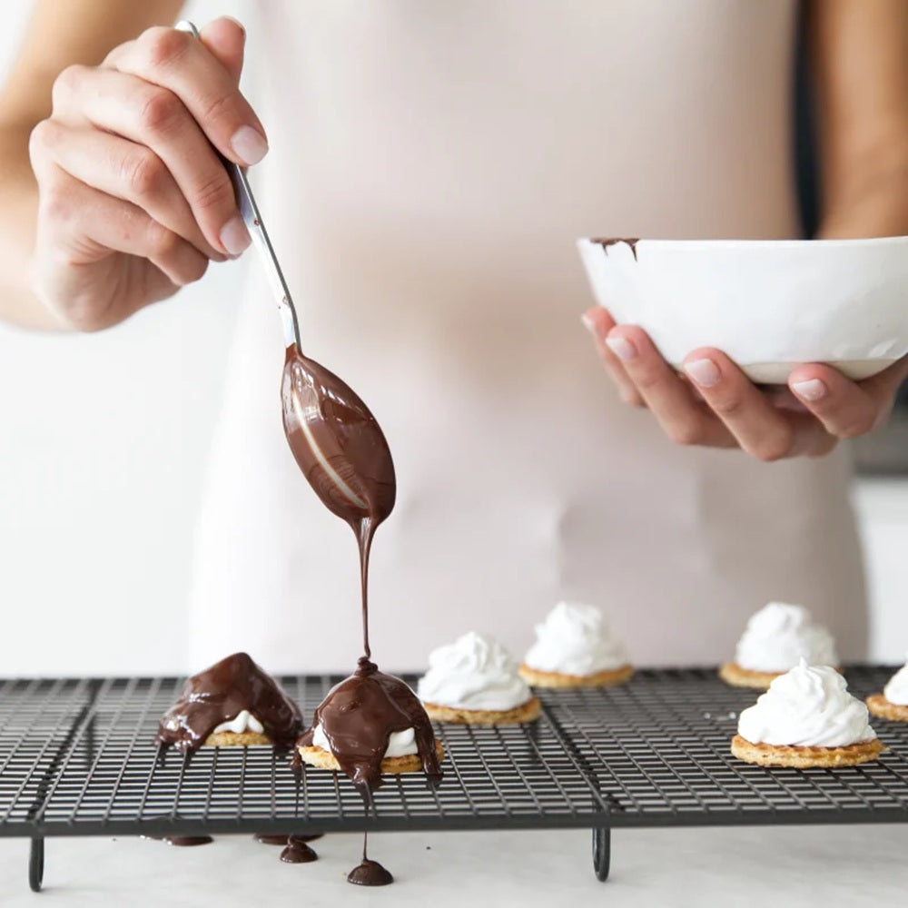 Person drizzling chocolate sauce over small desserts on a cooling rack.