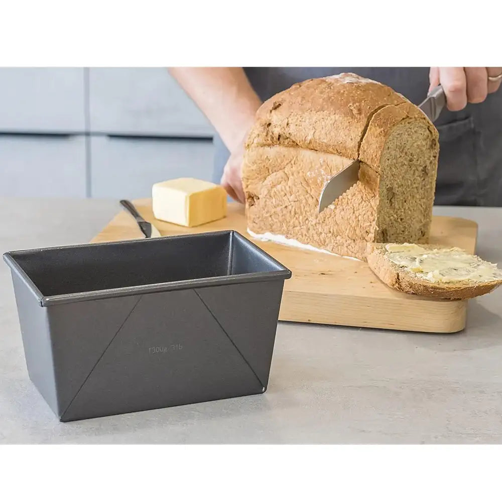 Person slicing bread on a wooden board with a loaf pan in the foreground.
