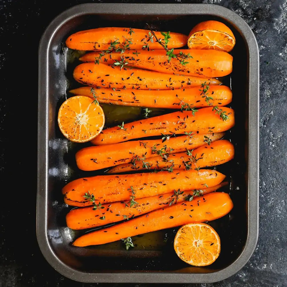 Roasted carrots with thyme and oranges in a baking tray on a dark surface