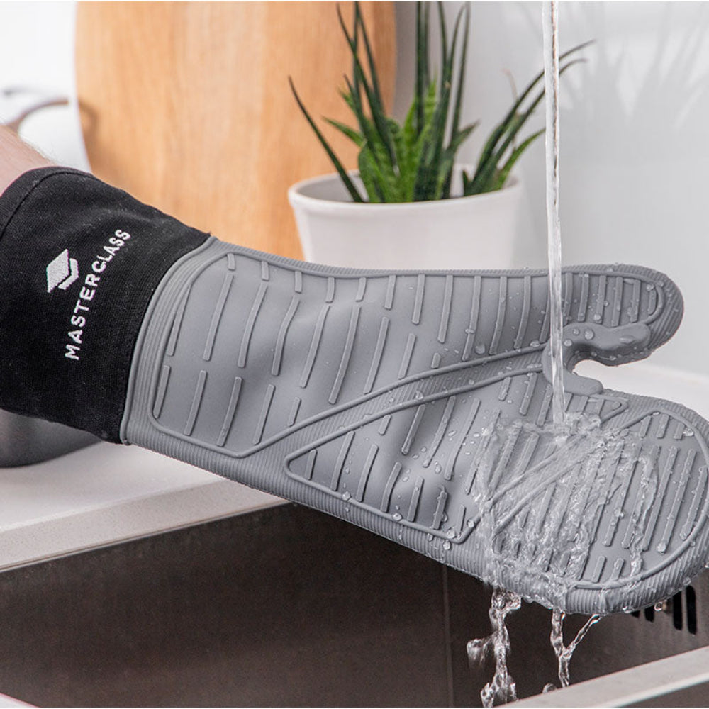 Gray silicone oven mitt with black cuff labeled 'Masterclass' being rinsed under running water.