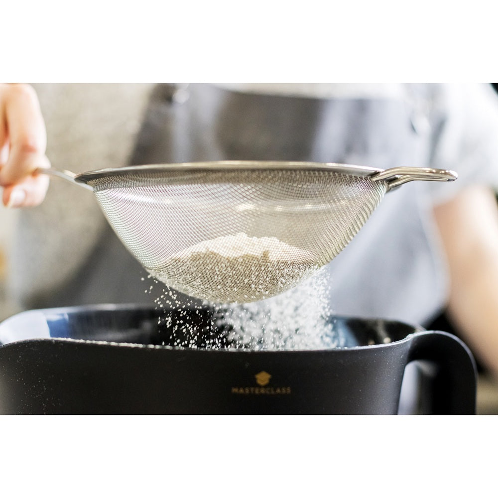 Person sifting flour into a black mixing bowl using a metal sieve.