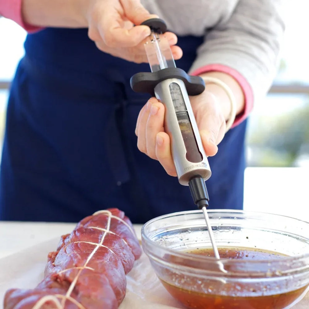 Person using a marinade injector to add flavor to a piece of meat with a bowl of marinade nearby.