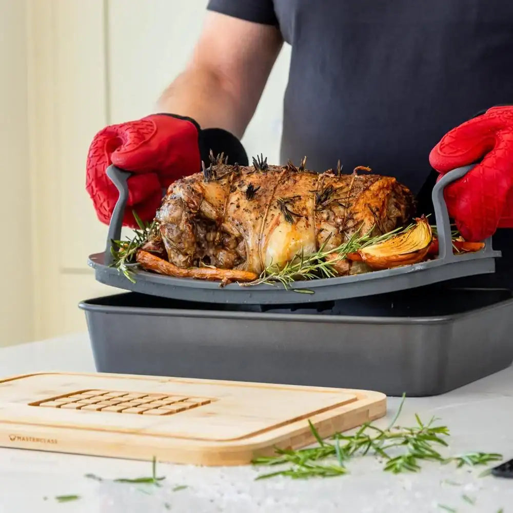 Person lifting a roasted dish with red gloves over a gray roasting pan on a light surface.
