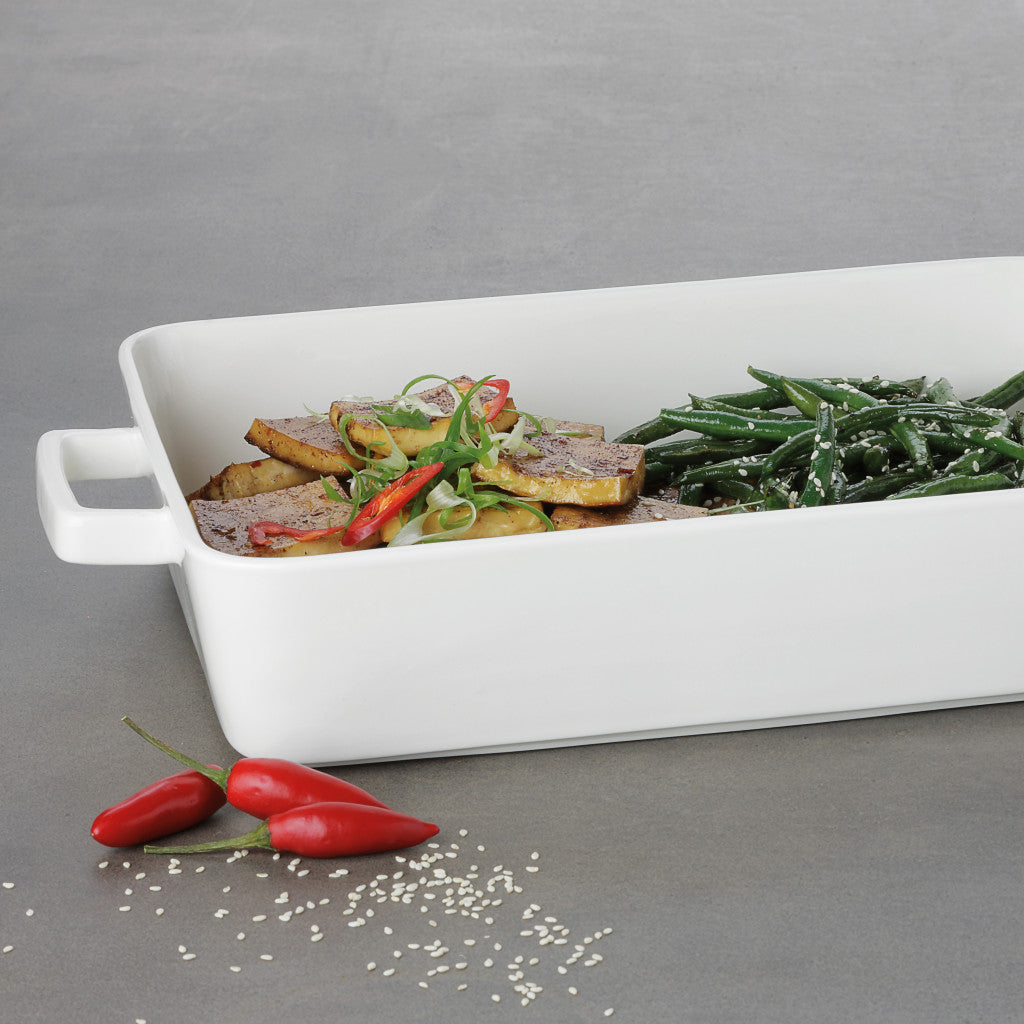 A white rectangular ceramic baking dish with side handles, shown filled with cooked food including green beans and sliced golden-brown pieces with red chili accents, on a gray surface with a few red chili peppers and sesame seeds in front.