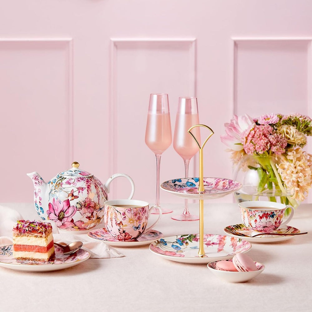 Table setting with floral teapot, cups, cake, and glasses on a pink background